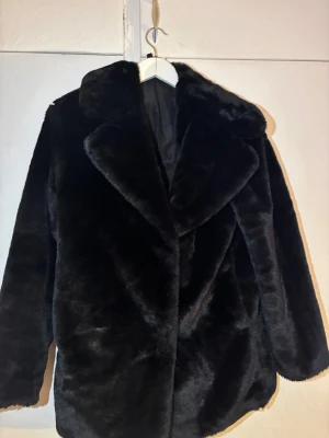 Svart pälsjacka  - Snygg svart pälsjacka med bred krage och klassisk rak passform. Jackan har en mjuk och glansig yta som ger en lyxig känsla. Perfekt för dig som vill ha en statement piece i garderoben. Fodrad insida för extra komfort och värme.