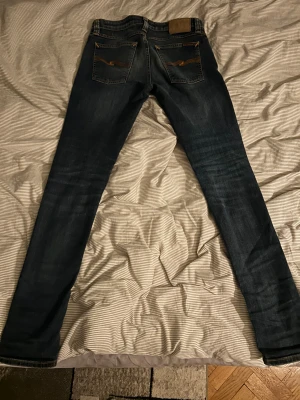 Mörkblå jeans från Nudie Jeans - Säljer ett par mörkblå  jeans från Nudie Jeans med klassiska orange sömmar och broderad bakficka. Jeansen har låg midja och är tillverkade i stretchig bomullsdenim för en tight passform. Perfekta för dig som gillar en smal siluett och stilren look. Hör av er vid frågor😁