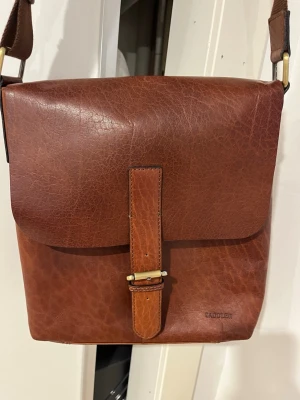 Brun axelväska i skinn från Saddler - Snygg brun axelväska från Saddler i äkta skinn med justerbar axelrem i textil. Väskan har ett klassiskt lock med spänne och en praktisk ytterficka på baksidan. Perfekt för dig som vill ha en stilren och tidlös väska med plats för det viktigaste.25x25cm