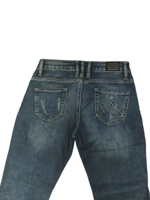 Philipp Plein blå jeans med dödskalle - Snygga blå jeans från Philipp Plein med slitningar och coola detaljer som dödskallebroderi på bakfickan och glittriga paljetter vid fickorna. Jeansen har en slim passform och är tillverkade i klassiskt denimtyg. Perfekt för dig som gillar edgy stil och unika detaljer. Storlek 30