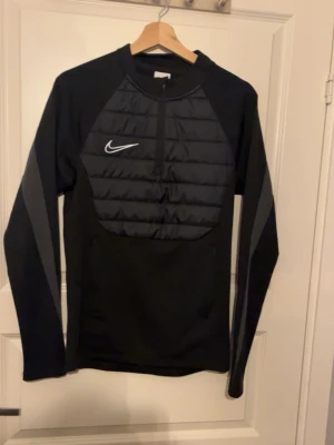 Svart Nike tröja med half zip - Säljer en svart långärmad tröja från Nike med quiltad front och half zip. Tröjan har en sportig look med grå detaljer på ärmarna och Nike-logga på bröstet. Perfekt för träning eller chill dagar. Materialet är syntet och polyester.