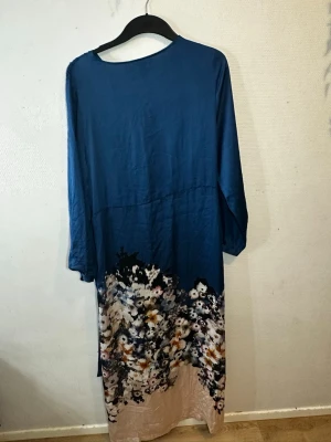 Blommig långklänning med v-ringning - Snygg långklänning i marinblått med v-ringad front och twistad detalj i midjan. Klänningen har långa ärmar och ett blommigt mönster i vitt, beige och blått nedtill. Materialet känns mjukt och har ett fint fall, perfekt för dig som gillar stilrena och romantiska plagg.