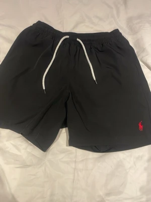 Svarta badshorts från Polo Ralph Lauren - Svarta badshorts från Polo Ralph Lauren med vit snörning i midjan och röd logga broderad framtill. Shortsen har elastisk midja, bakficka med knapp och innerfoder i mesh. Perfekta för strand eller poolhäng.