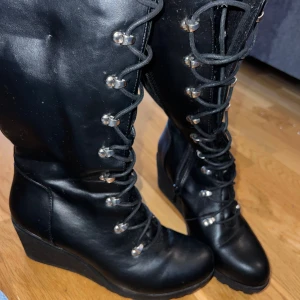 Svarta kängor med kilklack, storlek 40 - Säljer ett par svarta kängor i skinnimitation med snörning och dragkedja på insidan. Skorna har rund tå och en stabil kilklack som ger extra höjd utan att tumma på komforten. Metallhakar längs snörningen ger en cool detalj. Perfekta för dig som vill ha en edgy look. 6,5 cm hög klack 