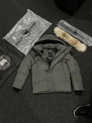 Grå pufferjacka från Canada Goose - Canada Goose pufferjacka i Grå med huva och avtagbar pälsdetalj. Jackan har dragkedja, knappar och märkets logga på ärmen. Insidan är fodrad och jackan är designad för att hålla dig varm under kalla dagar. Perfekt för dig som vill ha en stilren och funktionell vinterjacka.