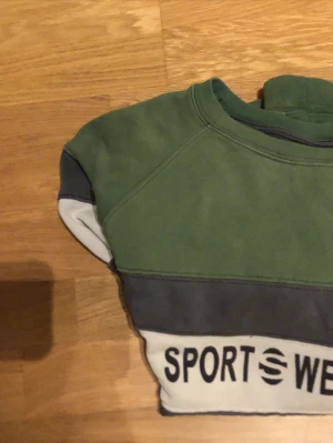 Grön och vit sweatshirt med tryck - Säljer en grön sweatshirt från Sportwear med breda vita och mörkgrå ränder samt trycket 'SPORTWEAR' på bröstet. Tröjan har rund hals och långa ärmar, perfekt för chill dagar. Materialet känns som mjuk bomull och passformen är normal.