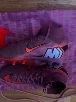 Rosa nike mercurial superfly 10 elite - Säljer ett par lila Nike Mercurial fotbollsskor med högt skaft och snörning. Skorna har en stor M-detalj på sidan, röda detaljer och sula med dobbar för gräsplan. Ovandelen är i syntetmaterial och de kommer med en matchande lila Nike-påse.