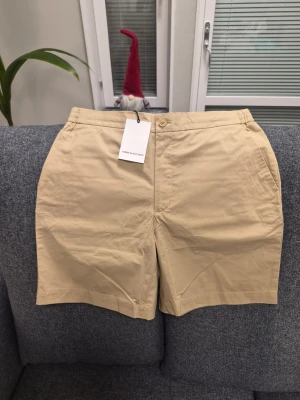 These Glory Days T Adel Poplin beiga shorts, storlek 52 - Snygga beige shorts från These Glory Days med rak passform och elastisk midja. De har både knapp och dragkedja framtill samt snörning för extra komfort. Tillverkade i mjuk bomull, perfekta för varma dagar. Fickor både fram och bak för praktisk förvaring.