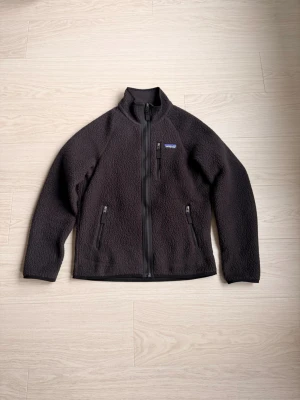 Svart fleecejacka från Patagonia - Mysig svart fleecejacka från Patagonia i storlek S. Jackan har hel dragkedja framtill, två sidofickor med dragkedja och en liten ståkrage. Perfekt för kyliga dagar och enkel att slänga på sig när du vill vara bekväm och varm. Bara använd 1 gång 