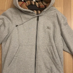 Grå huvtröja från Burberry - Snygg grå huvtröja från Burberry med dragkedja och klassiskt rutigt foder i huvan. Tröjan har en broderad logotyp på bröstet och praktiska fickor framtill. Perfekt för en stilren och bekväm look.