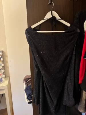 Svart glittrig kortklänning med öppen rygg - Svart glittrig kortklänning med snygg drapering och öppen rygg. Klänningen har halterneck med knyt i nacken och ett glittrande tyg som ger extra partyvibe. Perfekt för dig som vill sticka ut med en cool och elegant look.