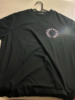 Svart Chrome Hearts t-shirt med broderi - Svart t-shirt från Chrome Hearts med stort broderat emblem och text på ryggen samt mindre broderi på bröstet. Klassisk rund hals och korta ärmar. Materialet är mjuk bomull och broderiet har vita detaljer med metallkors i mitten. Perfekt för dig som gillar streetwear och statement-plagg.