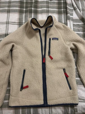 Patagonia fleece - Säljer en beige fleecejacka från Patagonia i storlek S. Jackan har mörkblå detaljer, hel dragkedja framtill och tre fickor med dragkedja och röda zip-pullers. Perfekt för kyliga dagar och har en mysig, fluffig känsla.