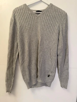Grå stickad tröja med v-ringning - Säljer en grå stickad tröja med v-ringad hals och kabelstickat mönster. Tröjan har långa ärmar och ribbade muddar vid ärmslut och nederkant. Perfekt för dig som gillar klassisk och enkel stil.