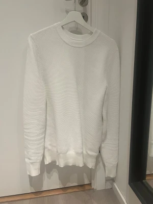 Vit stickad tröja från J.Lindeberg - Säljer en stilren vit stickad tröja från J.Lindeberg i storlek M. Tröjan har rund hals, ribbade muddar och ett diskret textur-mönster över hela ytan. Perfekt för dig som gillar enkel och clean design.