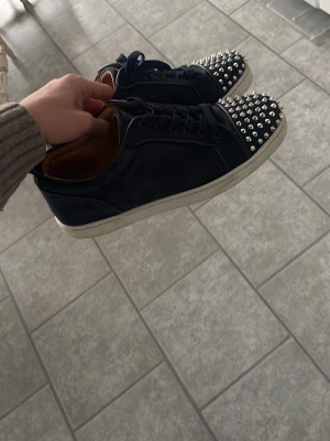 Mörkblå Christian Louboutin sneakers med nitar - Säljer ett par mörkblå sneakers från Christian Louboutin med vita sulor och coola silvriga nitar på tårna. Skorna har snörning och är tillverkade i skinn, vilket ger en lyxig känsla. Medföljer extra skosnören och tillbehör i originalpåse.