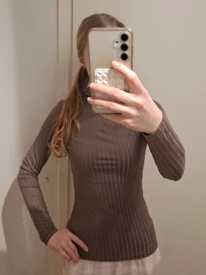ribbad långärmad tröja - Säljer en brun, ribbad långärmad topp med hög hals. Materialet är stretchigt och följsamt, vilket ger en slim och kroppsnära passform. Perfekt för dig som gillar en enkel men ändå stilren look. Inga synliga loggor eller märken.