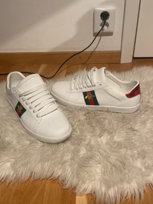 Gucci vita sneakers med bi-detalj - Säljer ett par vita Gucci sneakers i skinn med klassiska gröna och röda ränder på sidan och en guldbi broderad. Baksidan har metallicröd och metallicgrön detalj med Gucci-logga. Skorna har snörning och rund tå, och kommer med originalkartong och påse.