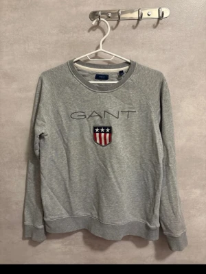 Grå sweatshirt från GANT - Säljer en grå sweatshirt från GANT med broderad logga och emblem på bröstet. Tröjan har rund hals, långa ärmar och ribbade muddar vid ärmslut och nederkant. Perfekt för en chill och stilren look.