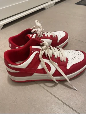 Nike Dunk Low röd och vit - Säljer ett par Nike Dunk Low sneakers i rött och vitt med klassisk snörning och perforerad tå. Skorna har en låg siluett och är tillverkade i skinn med vit sula och röda detaljer. Perfekt för dig som gillar streetwear och vill sticka ut med snygga färger.