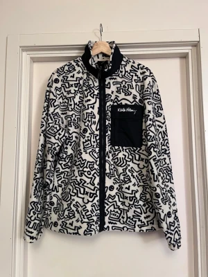 Keith Haring x H&M Fleecejacka  - Unik fleecejacka från Keith Haring x H&M kollektionen med svartvitt grafiskt mönster och svart bröstficka med logga. Jackan har hel dragkedja, hög krage och är långärmad. Perfekt för dig som vill sticka ut med streetstyle och konstnärlig vibe. Stor relaxed fit, S är som en L. Mycket bra skick, som ny! Sälj på flera sidor, först till kvarn. 