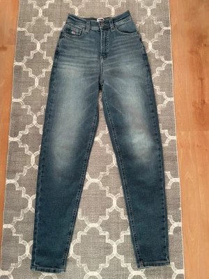 Tommy Jeans Mom Slim Ultra High - Snygga blå mom jeans från Tommy Jeans med ultra hög midja och slim passform. Klassisk femficksmodell med logga på lilla fickan och knapp med märkets namn. Tillverkade i mjukt denim med 94% bomull, 4% polyester och 2% elastan för lite stretch. Storlek 25/34. Använda en gång! Stretchiga med hög midja.