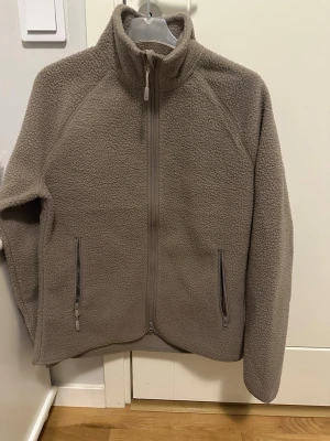 Beige fleecejacka från Lager 157 - Mysig beige fleecejacka från Lager 157 med hög krage och hel dragkedja framtill. Jackan har två praktiska fickor med dragkedja och en skön, teddy-liknande yta. Perfekt för kyliga dagar och enkel att matcha med din vardagsstil.