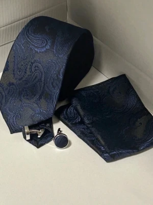 Mörkblå paisleymönstrad slipsset 1234 - Säljer ett stilrent set med mörkblå slips, matchande näsduk och manschettknappar. Slipsen har ett elegant paisleymönster i blå toner som ger en lyxig känsla. Perfekt för dig som vill lyfta din outfit med klassiska accessoarer.