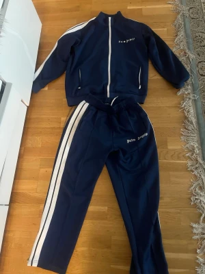 Palm Angels blå mjukisbyxor M - Säljer ett par snygga blå mjukisbyxor från Palm Angels i storlek M. Byxorna har vita ränder längs sidorna, elastisk midja med snörning och Palm Angels-logga på benet. Materialet är mjukt och skönt, perfekt för chill eller träning.