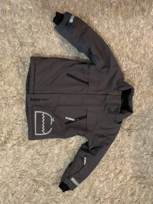 Grå vindjacka från Kaxs Proxtec - Grå vindjacka från Kaxs Proxtec med hög krage, svarta dragkedjor och reflexdetaljer på ärmarna. Jackan har två snedställda fickor med dragkedja och ett stilrent tryck på ena sidan. Materialet är slitstark polyester, perfekt för blåsiga dagar.