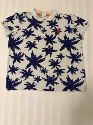 Vit och marinblå polotopp från USPA - Säljer en fräsch polotopp från USPA i vit med marinblå palmtryck över hela toppen. Klassisk krage, två knappar framtill och orange broderad logga på bröstet. Perfekt för sommarens chill dagar. Materialet är mjuk bomull.