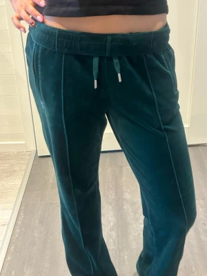 Mörkgröna mjukisbyxor Juicy Couture - Säljer ett par mörkgröna mjukisbyxor från Juicy Couture i mjuk sammet med strassdetaljer. Byxorna har snörning i midjan, raka ben och Juicy-logga i strass på baksidan samt ett litet emblem framtill. Perfekt för dig som gillar glam och comfy vibes.