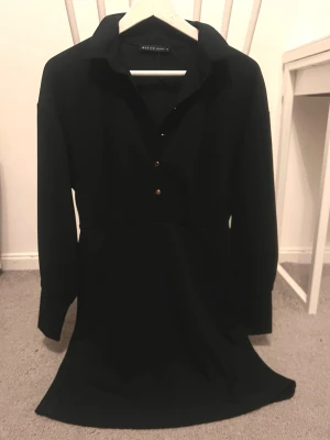 Svart skjortklänning - Stilren svart skjortklänning med klassisk krage och knappar framtill. Klänningen har lång ärm och en kortare längd som ger en modern look. Perfekt för dig som gillar minimalistisk stil och vill ha något enkelt men snyggt.