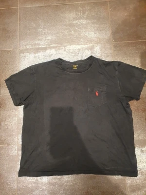 Svart t-shirt från Polo Ralph Lauren XL - Svart t-shirt från Polo Ralph Lauren i storlek XL. Klassisk design med korta ärmar och rund hals. På bröstet finns en liten ficka med röd broderad Polo-logga. 