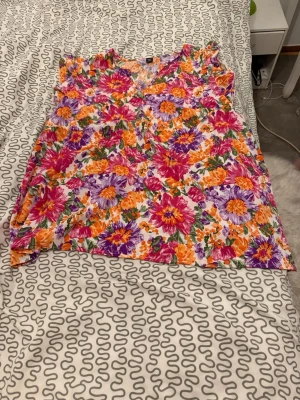 Blommig blus med v-ringning - Säljer en färgglad blus med v-ringning och blommigt mönster i lila, orange, rosa och grönt. Blusen är ärmlös och har en luftig passform, perfekt för varma dagar. Materialet känns lätt och bekvämt mot huden.