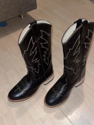 Svarta cowboy boots - Säljer ett par svarta cowboy boots med vit  brodering och ljus insida. Bootsens ovansida har snygga mönster och kontrastsömmar, och sulan är i ljust material. Perfekta för dig som vill sticka ut med westernstil. Välbevarat skick 