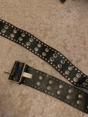 Svart nitbälte från Saddler - Säljer ett svart bälte från Saddler med bred design och massor av silvriga nitar och hål längs hela bandet. Bältet är gjort i skinn och har en rejäl metallspänne som ger en cool och edgy vibe. Perfekt för att lyfta din outfit.