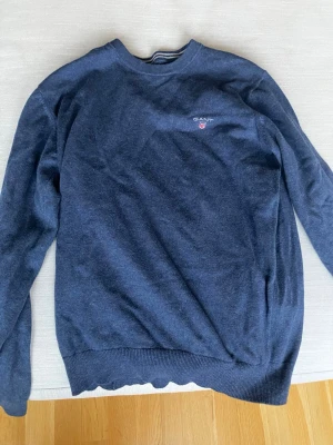 Mörkblå stickad tröja från GANT - Snygg mörkblå stickad tröja från GANT i storlek S. Tröjan har rund hals med ribbad kant och diskret logga broderad på bröstet. Perfekt för dig som gillar klassisk och enkel stil. Passar till jeans eller chinos för en chill look.