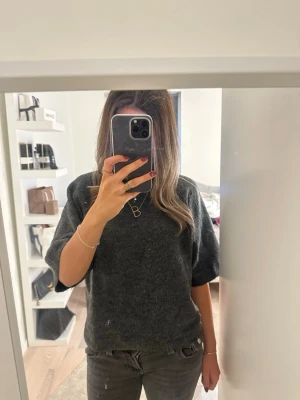 Mörkgrå stickad tröja från 157 - Mysig mörkgrå stickad tröja från 157 med avslappnad passform och halvlånga ärmar. Perfekt till höst och vinter, enkel att styla med jeans eller kjol. Ribbad kant vid halsen för extra detalj.