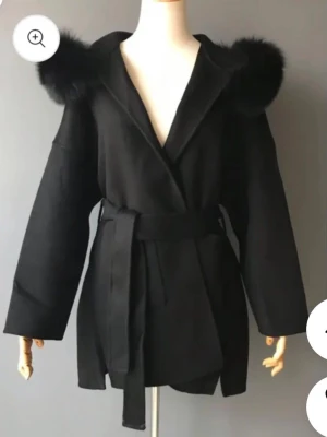 Kapp från FUR Stockholm  - Säljer en svart kappa med stor päls på luvan och knytskärp i midjan. Pälsen är avtagbar. Jackan har en omlottmodell och vida ärmar. Funkar både på vinter & vår. Nypris ca 2 000kr. 