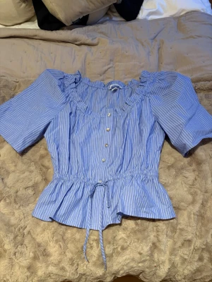 Blå randig blus från H&M - Säljer en blå blus med vita ränder från H&M i storlek S. Blusen har korta puffärmar, knappar framtill och en söt volang vid halsen. Midjan är markerad med snörning och blusen har en lätt croppad passform. Perfekt för vår och sommar! Pris kan diskuteras!!💞