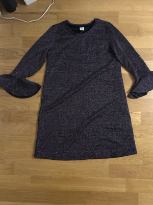 Glittrig svart klänning - Snygg svart glittrig klänning från Lindex med långärmade trumpetärmar. Klänningen har en rak passform och är tillverkad i ett glittrigt syntetmaterial som ger en cool look. Perfekt för dig som gillar att sticka ut med lite extra glitter. Perfekt för nyår eller fest.