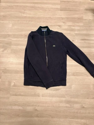 Marinblå ziptröja från Lacoste - Säljer en marinblå ziptröja från Lacoste med klassisk ståkrage och silverfärgad dragkedja. Tröjan har lång ärm, två fickor framtill och den ikoniska gröna krokodilloggan på bröstet. Skön passform och perfekt till lager på lager under höst och vår.