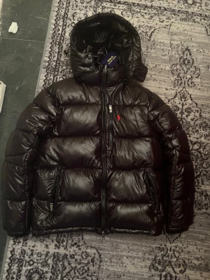 Svart pufferjacka från Polo Ralph Lauren - Säljer en svart pufferjacka från Polo Ralph Lauren med blank finish och röd broderad logga på bröstet. Jackan har huva, flera dragkedjeförsedda fickor och en schysst oversized look. Perfekt för kalla dagar när du vill vara både varm och stilren.