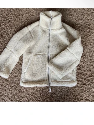 Vit teddyjacka med dragkedja - Mysig vit teddyjacka med hög krage och hel dragkedja framtill. Jackan har två stora fickor och är tillverkad i fluffigt fleece-material som håller dig varm. Perfekt för kyliga dagar och har en enkel, stilren look.