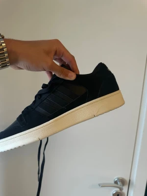 Svarta Adidas sneakers med vit sula - Säljer ett par svarta Adidas sneakers med klassiska tre ränder på sidan och beige sula. Skorna har snörning och är tillverkade i mocka och syntet. Perfekta för dig som gillar stilrena och tidlösa sneakers.