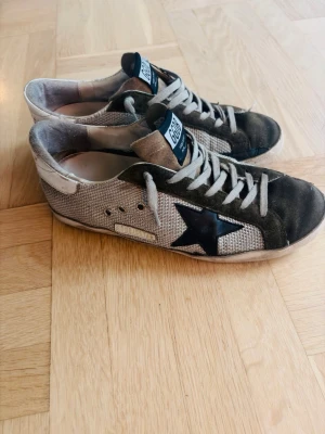 Golden Goose sneakers  - Säljer ett par Golden Goose sneakers i beige och svart med GGDB-logga på plösen och en ikonisk svart stjärna på sidan. Skorna har snörning, rund tå och platt sula. Ovandelen är i textil och mocka, med vita detaljer på hälen och slitna kanter för en cool vintage-look. Sulan är lite lös men inget som man inte kan limma. Skriv gärna om du har någon fråga! 