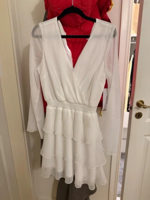 Vit omlottklänning med volanger - Säljer en superfin vit kortklänning med omlottdesign och v-ringning. Klänningen har långa ärmar, smock i midjan och flera lager volanger i kjolen för en flowy look. Perfekt till student! Prislapp fof kvar då den aldrig använts.