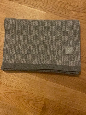 Grå rutig set från Lv ÄKTA - Snygg grå halsduk från Lv med klassiskt rutmönster och diskret LV-logga i ena hörnet. Halsduken är mjuk och stilren, perfekt för att lyfta din outfit. Materialet känns som ullblandning och färgen är en mix av ljus- och mörkgrå toner.