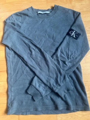 Grå långärmad tröja Calvin Klein - Snygg grå stickad långärmad tröja från Calvin Klein Jeans med ribbade muddar och en svart CK-logga på ärmen. Tröjan har rund halsringning och är tillverkad i ett mjukt, strukturerat material som känns bekvämt mot huden.